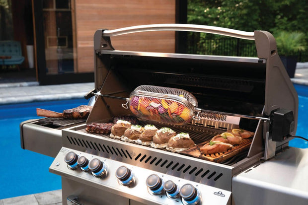 Napoleon gasbarbecue met open deksel, beladen met gegrilde steaks, gevulde aardappelen, zalm op een cederhouten plank en groenten in een rotisserie-mand, naast een buitenzwembad